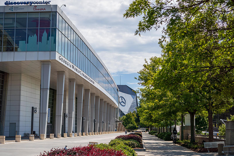 Exterior view of the Georgia World Congress Center building with columns along a sidewalk lined with green trees on a partly cloudy day.
