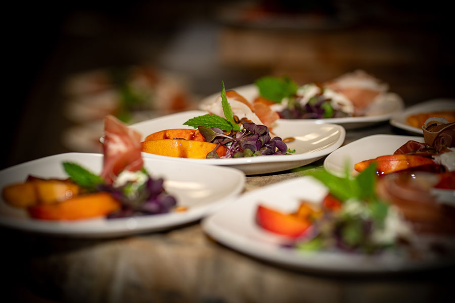 Close-up of multiple white plates with sliced peaches, green mint leaves, purple microgreens, and thin slices of prosciutto on a rustic wooden table.