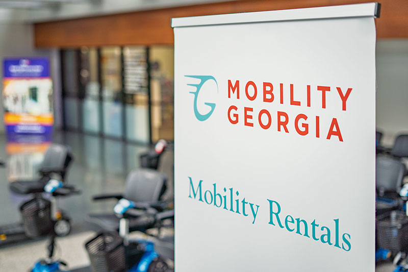 Sign with Mobility Georgia logo and text 'Mobility Rentals' in a showroom with mobility scooters in the background.