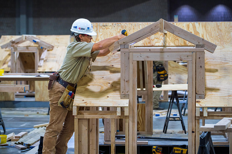 Construction worker wearing a white hard hat assembling a wooden frame structure indoors.