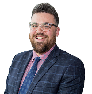Smiling man with glasses, beard, and curly hair wearing a blue checkered suit, pink shirt, and blue tie in a bright indoor setting.