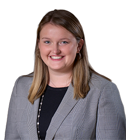 Smiling woman with shoulder-length blonde hair wearing a gray checkered blazer and black top against a blurred indoor background.