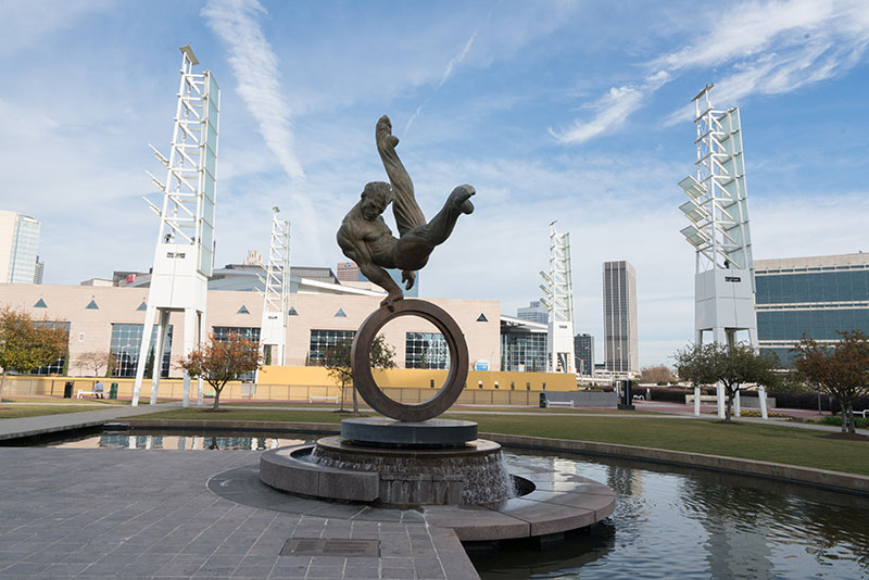 Sculpture of a man performing a dynamic acrobatic move on top of a circular ring above a water fountain, set in an urban plaza with modern buildings and towers in the background.