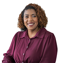 Portrait of a smiling woman with curly hair wearing a black blazer and silver necklace in an indoor setting.