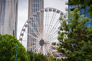 Large Ferris wheel partially obscured by green trees and surrounded by tall modern buildings in an urban setting.