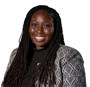 Smiling woman with long dreadlocks wearing a patterned blazer and black top against a blurred dark background.